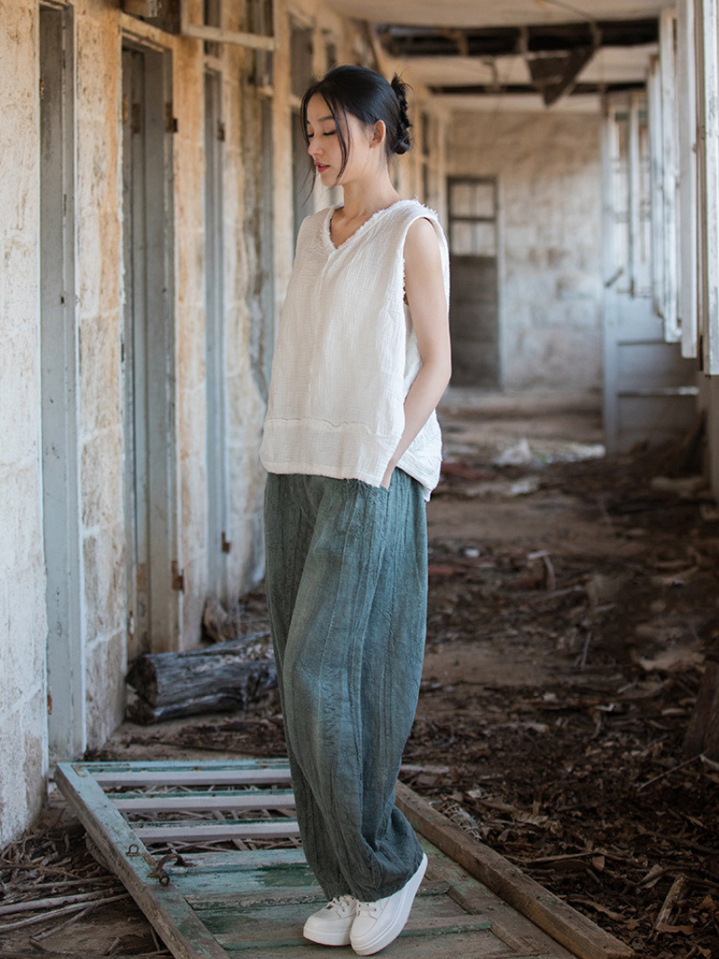 Woman standing in an abandoned building wearing a white sleeveless top and green pants.